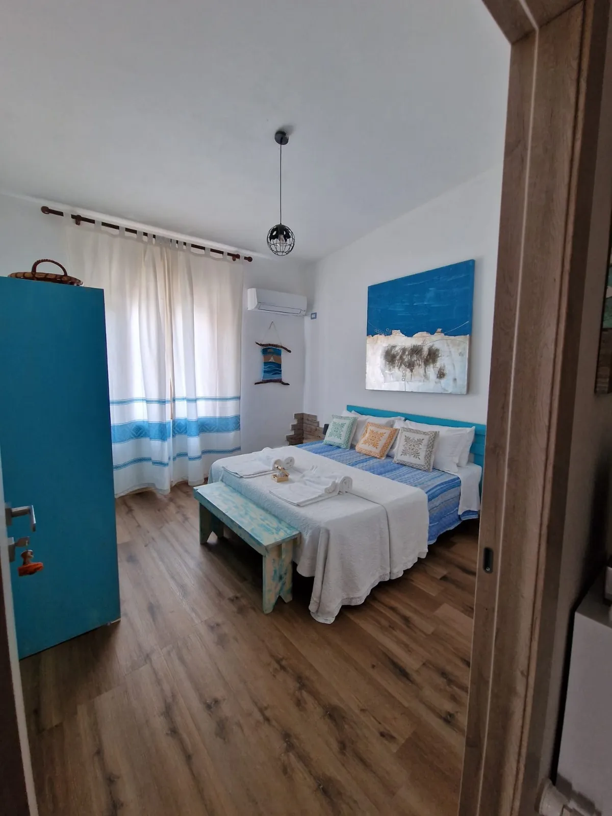Camera Cala Luna: letto con copriletto a righe blu, testiera turchese, quadro astratto del mare, tende sarde e panchetta in legno turchese