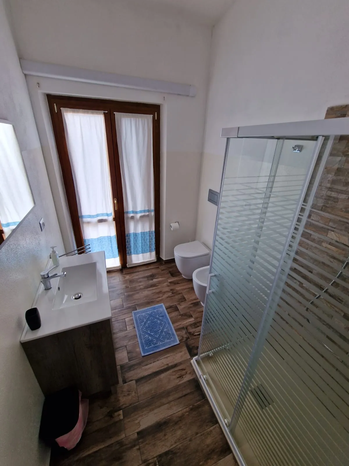 Bagno privato della camera Cala Luna con box doccia, parete in pietra, pavimento effetto legno e porta-finestra con tende sarde