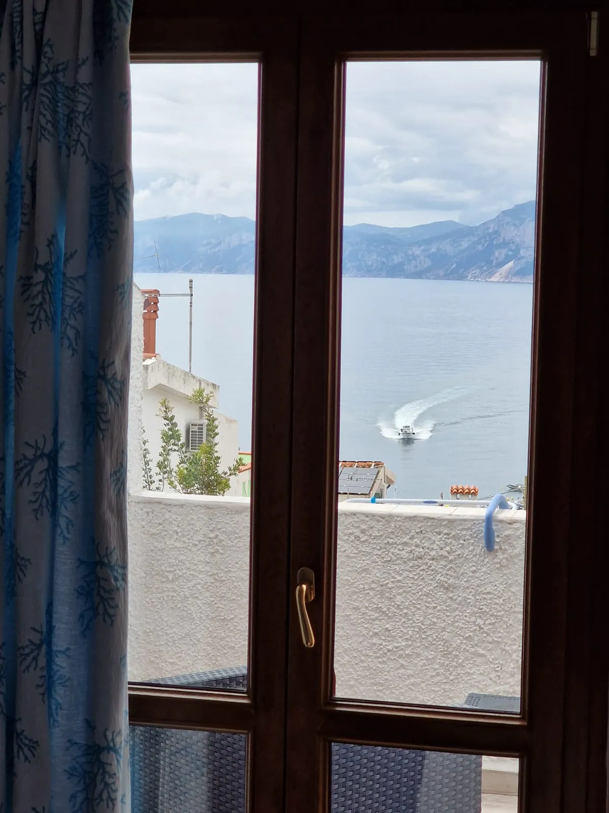 Porta-finestra della camera Cala Luna con tenda a motivo corallo e vista sul mare con una barca che lascia la scia