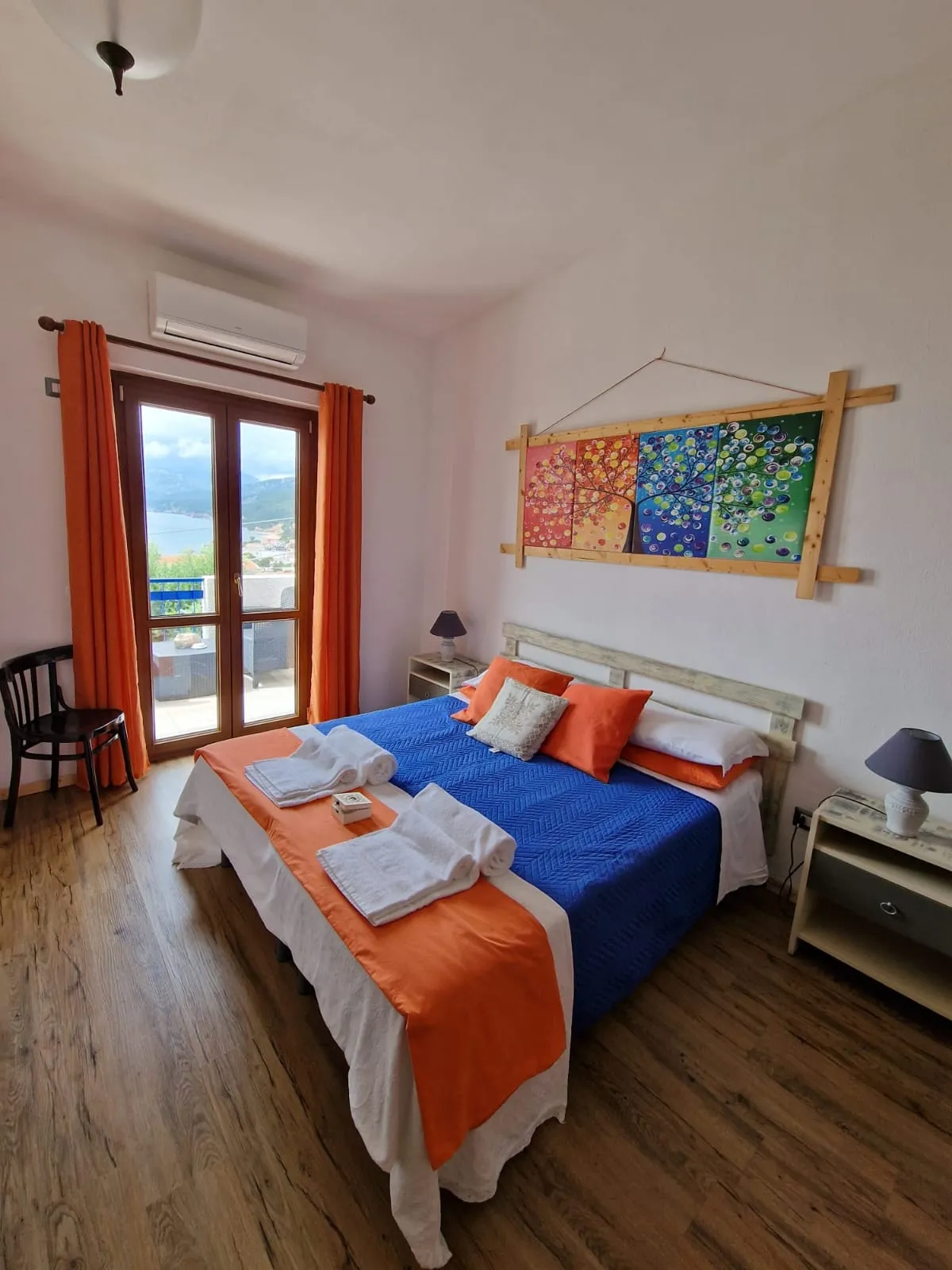 Camera Cala Goloritzè: copriletto blu trapuntato con runner arancione, quadro colorato delle quattro stagioni e porta-finestra sul balcone vista mare