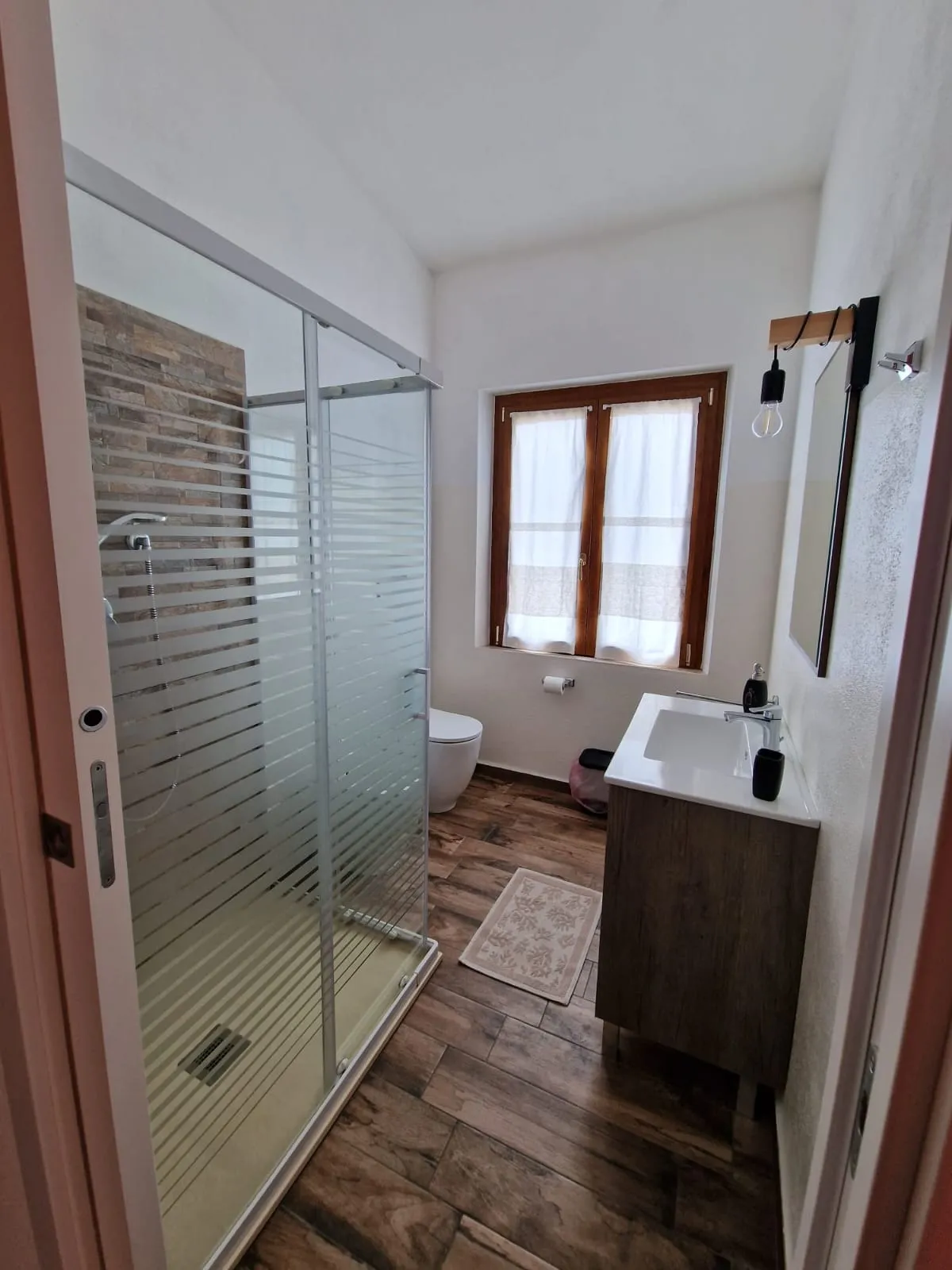 Bagno privato della camera Cala Goloritzè con box doccia, parete effetto pietra e finestra in legno con tende bianche