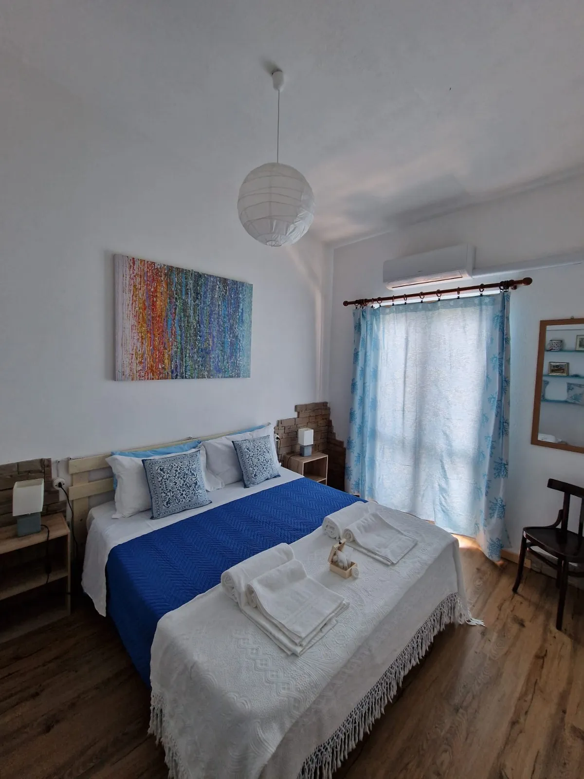 Camera Monte Litty: copriletto blu con coperta bianca a frangia, quadro a righe verticali multicolore e lampada a sfera di carta